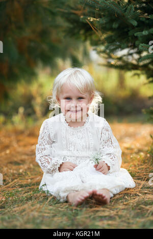 Eine schöne 2-jähriges Mädchen Kind ist draußen zu sitzen, Lächeln unter den Pinien bei Sonnenuntergang auf einem späten Sommerabend. Stockfoto