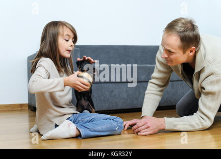 Vater und Tochter spielen mit dem Welpen auf dem Boden im Zimmer. Das Mädchen ist die Kugel geben dem Hund. Stockfoto