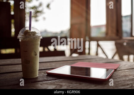 Ein Glas mit Smoothies und eine Tablette auf einem hölzernen Tisch auf der Veranda von einem Cafe. Stockfoto