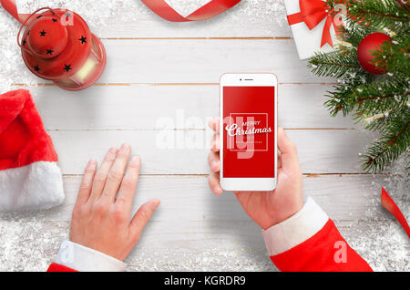 Santa mit Handy am Schreibtisch. Weihnachtsbaum mit Dekorationen, Geschenke und Laterne auf weißem Holzschreibtisch. Ansicht von oben. Stockfoto