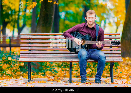 Junger Mann sitzt auf der Bank und spielt Akustikgitarre im City Park Stockfoto