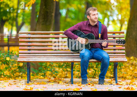 Junger Mann sitzt auf der Bank und spielt Akustikgitarre im City Park Stockfoto