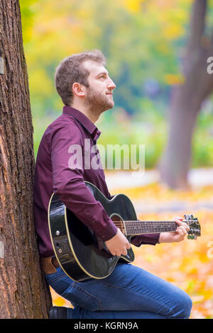 Junge Mann sitzt und spielt Akustikgitarre im City Park. Fokus auf seinem Auge! Stockfoto