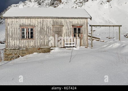 Schnee bedeckt völlig ruiniert - verlassen - Farbe verblasst Holzhütte mit Holzrahmen der Schaukel - Fuß des schneebedeckten Mount hustinden neben e10 European Road Stockfoto