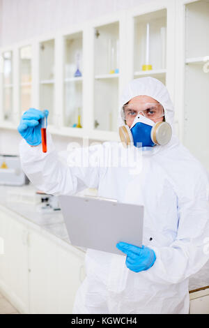 Mann in Schutzkleidung, Handschuhe, Schutzanzug und Gasmaske holding Dokument und Kolben mit roten Flüssigkeit Stockfoto