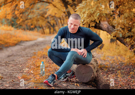 Eine junge sportliche Jogger in Schwarz enganliegende sportswears und Turnschuhe sitzt auf einem anmelden, fühlt sich eine starke Schmerzen im Muskel nach Einzwängen auf einem autumna Stockfoto