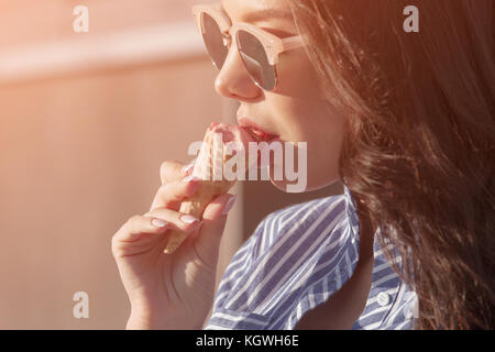 Junge, schöne Brünette Frau essen rosa Eis in der Waffel Horn. Sie Brille trägt mit dunkler Brille, und ein Hemd in einem blauen und weißen Streifen. cl Stockfoto