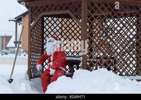 Müde weihnachtsmann sitzt auf Holzstuhl nach der Arbeit von Schneeräumung vor dem Holzhaus Stockfoto