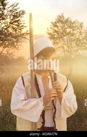 Junge Mann in der traditionellen albanischen Kostüm spielt Flöte vor der Natur Wiese und Abenddämmerung Stockfoto