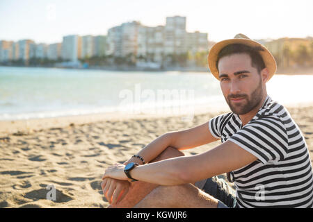 Porträt der jungen gutaussehenden Mann in Hut sitzen am Strand. Stockfoto