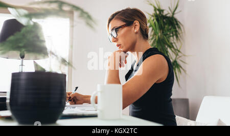 Frau mit Brille sitzt an ihrem Schreibtisch im Büro. Geschäftsfrau, die sich einen Laptop anschaut und Notizen schreibt. Stockfoto