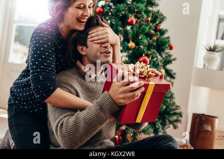 Ein paar glückliche Momente verbringen während Weihnachten zu Hause feiern. Lächelnden jungen Frau die Augen ihres Partners während über Weihnachten gif Übergabe Stockfoto