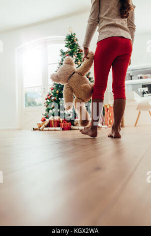 Niedrige Abschnitt der jungen Mädchen, dass Teddybären zu Fuß in Richtung Weihnachtsbaum. Geschmückten Weihnachtsbaum und Geschenke im Wohnzimmer angeordnet. Stockfoto