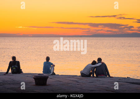 Menschen Meer romantisch, Rückansicht von jungen Menschen sitzen auf einer Hafenmauer und beobachten einen Sonnenuntergang an der Adria, Triest, Italien. Stockfoto