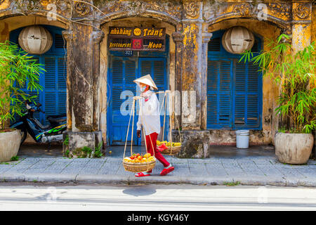 Hoi An, Vietnam; Street Scene von der Altstadt entfernt. Junge Frau, die ihre Früchte tragen auf der Schulter Bambusstab Stockfoto