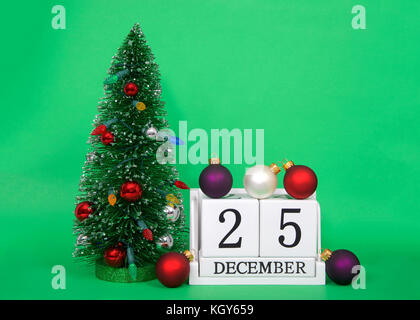 Holzblöcke Übersicht Kalender Datum des 25. Dezember, Weihnachten, sitzt auf einem grünen Hintergrund mit kleinen dekoriert Xmas Tree daneben, mehrere Stray orn Stockfoto