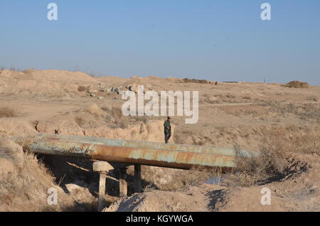 Alter Mann mit Jacke und Kappe gehen über ein Rohr im Boden über einen kleinen Fluss in der Wüste in Usbekistan. Stockfoto