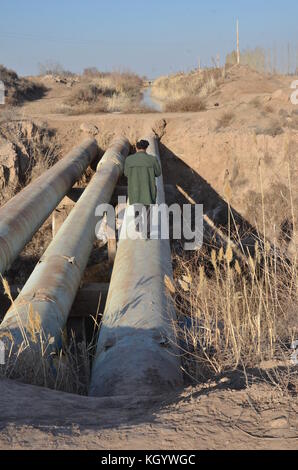 Alter Mann mit Jacke und Kappe gehen über ein Rohr im Boden über einen kleinen Fluss in der Wüste in Usbekistan. Stockfoto