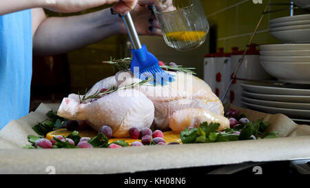 Weibliche Hände Bürsten ganze rohe Huhn mit Braten Soße in den Bräter mit orangen Preiselbeeren und Kräutern im Ofen Fach. Stockfoto