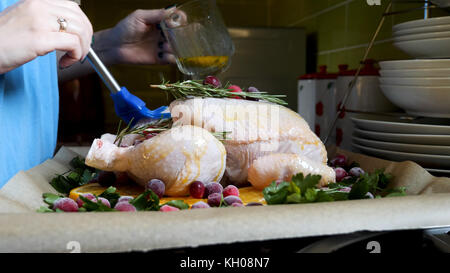 Weibliche Hände Bürsten ganze rohe Huhn mit Braten Soße in den Bräter mit orangen Preiselbeeren und Kräutern im Ofen Fach. Stockfoto