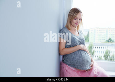 Es ist ein Mädchen! Schwangere Frau saß auf dem Fensterbrett in einem rosa Rock und ein T-Shirt. Stockfoto