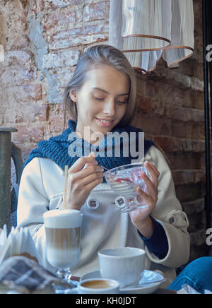 Eine charmante junge Dunkelhaarige ruht in einem gemütlichen Cafe. Kaffee wird auf Ihrem Tisch. Das Mädchen genießt den Geschmack von Eis. Stockfoto