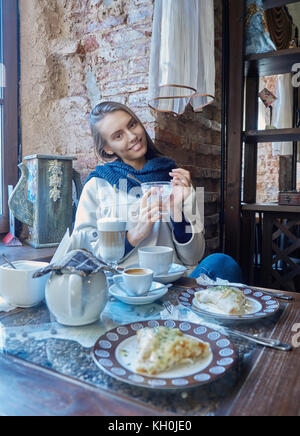 Eine charmante junge Dunkelhaarige ruht in einem gemütlichen Cafe. Kaffee wird auf Ihrem Tisch. Das Mädchen genießt den Geschmack von Eis. Stockfoto