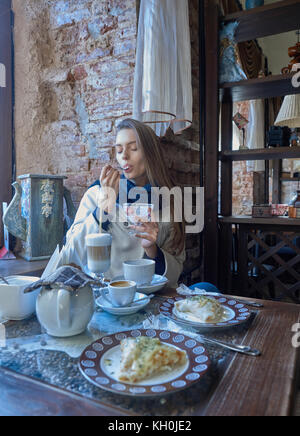 Eine charmante junge Dunkelhaarige ruht in einem gemütlichen Cafe. Kaffee wird auf Ihrem Tisch. Das Mädchen genießt den Geschmack von Eis. Stockfoto