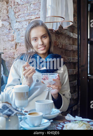 Eine charmante junge Dunkelhaarige ruht in einem gemütlichen Cafe. Kaffee wird auf Ihrem Tisch. Das Mädchen genießt den Geschmack von Eis. Stockfoto