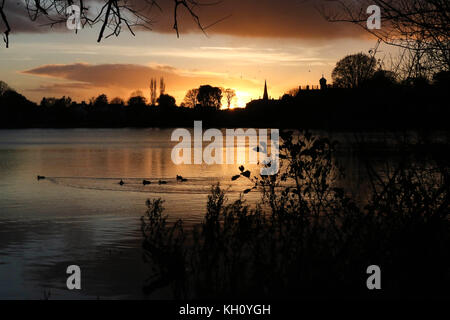 Lurgan Park See, Nordirland, Großbritannien. 12. November 2017. Ein blustery Tag mit kalten Nord-westlichen Winden und Duschen endet mit Clearing Skies und fallende Temperaturen. Frost zu rechnen ist, möglicherweise als niedrig zu-4C in ländlichen Gebieten, wie die Sonne auf der linken Seite von Shankill Pfarrkirche und Brownlow House (rechts). Quelle: David Hunter/Alamy Leben Nachrichten. Stockfoto