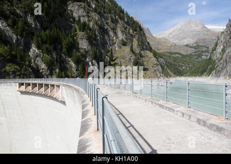 Blick von oben auf dem Gibidum Damm in Richtung zu seinem abflußkanäle mit dem Bügeleisen Schutzzaun im Vordergrund und eine alpine Landschaft im Hintergrund. Stockfoto