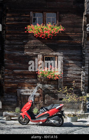 BLATTEN, SCHWEIZ - SEPT. 25, 2017: Ein roter Motorroller vor der Fassade eines alten Chalet aus Holz mit Blumen in Blumenkästen durch das Sonnenlicht. Stockfoto