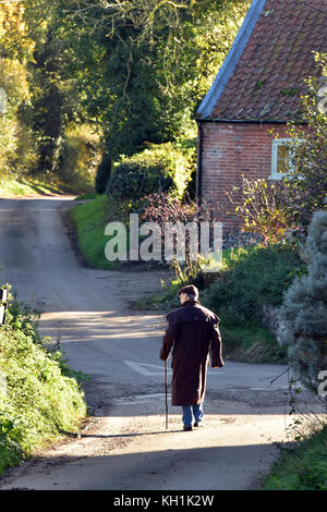 Ein Land gentleman Spaziergang durch ein Dorf entlang einer malerischen Lane in Norfolk an einem strahlenden Herbsttag mit einem Stick und trägt ein flacher Deckel. Stockfoto