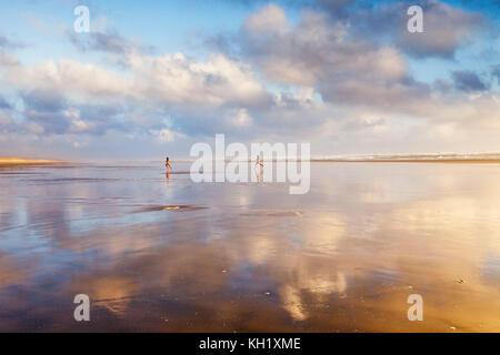 Junge Paare, die bei Sonnenuntergang über Ninety Mile Beach, Northland, Neuseeland, laufen. Stockfoto