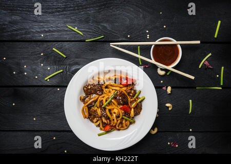 Udon fry Nudeln mit Fleisch und Gemüse in einer weißen Platte auf schwarzem Hintergrund Holz umrühren. mit Stäbchen und Soße. Ansicht von oben. Stockfoto