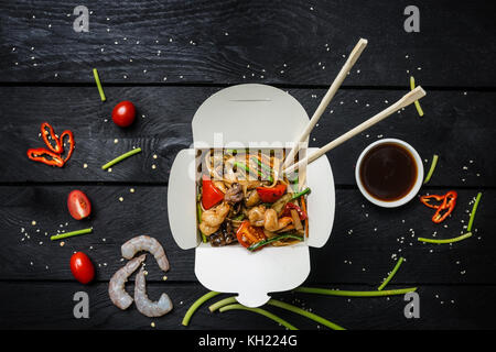 Udon fry Nudeln mit Meeresfrüchten und Gemüse in einer Box auf schwarzem Hintergrund Holz umrühren. mit Stäbchen und Soße. Ansicht von oben. Stockfoto