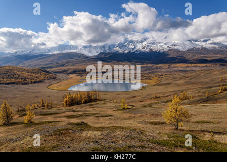 Helle malerischen Herbst Landschaft mit gelben Lärchen, einem wunderschönen See in der Steppe und der Berge, bedeckt mit Schnee auf dem Hintergrund des blauen Himmels und Stockfoto