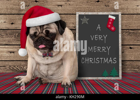 Grumpy mops Welpe Hund mit roten santa hat neben Blackboard Schild mit Text sehr frohe Weihnachten sitzen, auf hölzernen Hintergrund Stockfoto