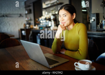 Junge asiatische Mädchen mit Laptop schauen neugierig Video im Cafe, das von Tasse trinken Stockfoto