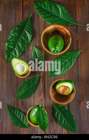 Grün roh reif und ganze avocado Früchte mit Stein liegen in Holzschalen durch Blätter auf einem braunen Tisch umgeben. Flach. top View Stockfoto