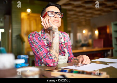Portrait von Kreative junge Frau mit Tattoos auf dem Arm am Tisch sitzen in Künstler workshop Suchen nachdenklich nach Inspiration suchen Stockfoto