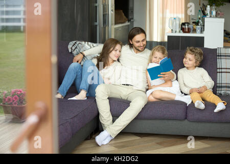 Glückliche Familie mit Kindern, die ein Buch auf dem Sofa halten, Vater umschließt die Mutter und liest Märchen für Kinder, die sich auf der Couch entspannen, Eltern und Sohn Tochter Stockfoto