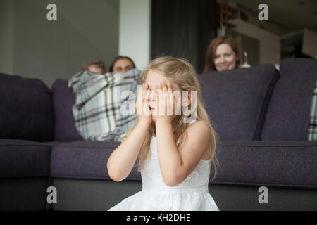 Kleines Mädchen, schließen die Augen, das Gesicht mit den Händen spielen Verstecken und Spiel Suchen mit Eltern und Bruder versteckt sich hinter Sofa peeking in Wohnzimmer, h Stockfoto