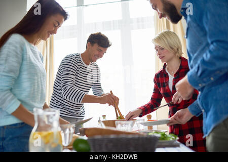Gruppe von jungen Menschen bereiten das Abendessen für festliche Feier steht am grossen Tisch essen und glücklich lächelnd Stockfoto