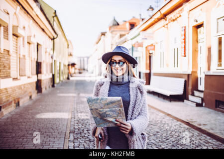 Glückliche Frau Tourist mit Karte, während auf der Straße stand, trendige Frau Reisenden mit Lächeln lesen geographischen Atlas während in der Stadt unterwegs.co Stockfoto