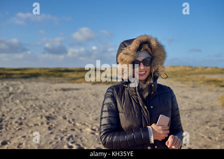 Trendy Frau in einem warmen Herbst Outfit mit Sonnenbrille, stehend auf einem Strand bei Sonnenuntergang wirft einen langen Schatten über den Sand an der Seite mit einem Ha suchen Stockfoto