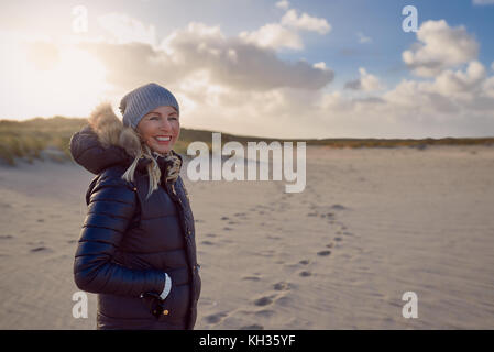 Trendy Frau in einem warmen Herbst Outfit stehen auf einem Strand bei Sonnenuntergang wirft einen langen Schatten über den Sand auf der Seite mit einem glücklichen Lächeln Stockfoto