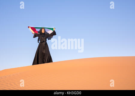 Schöne Frau in den nationalen UAE Kleid abaya Holding die Vereinigten Arabischen Emirate Flagge in der Wüste. Stockfoto