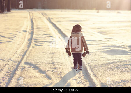 Ein kleiner Junge gekleidet Schaffellmantel und Kappe gehen auf Winter, verschneiten Wald Straße. Rückansicht. Spuren der Räder, das Auto auf der schneebedeckten Straße Stockfoto