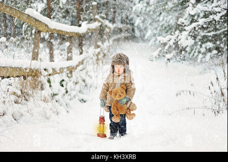 Eine kleine traurige, verlorene Junge stehend auf den Winter, verschneiten Wald Straße und halten Teddybären und Öl lampe Stockfoto